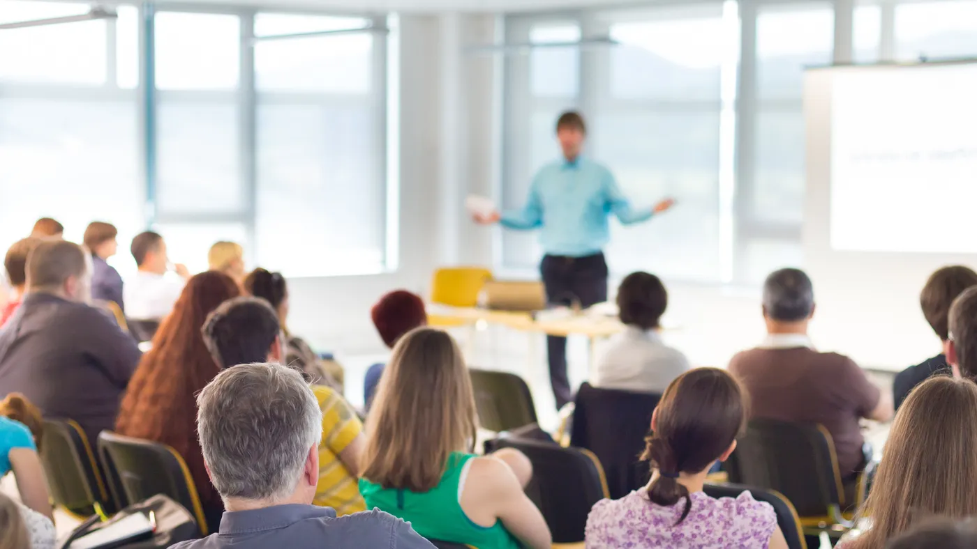 People seated at a public school-board meeting, facing a presenter