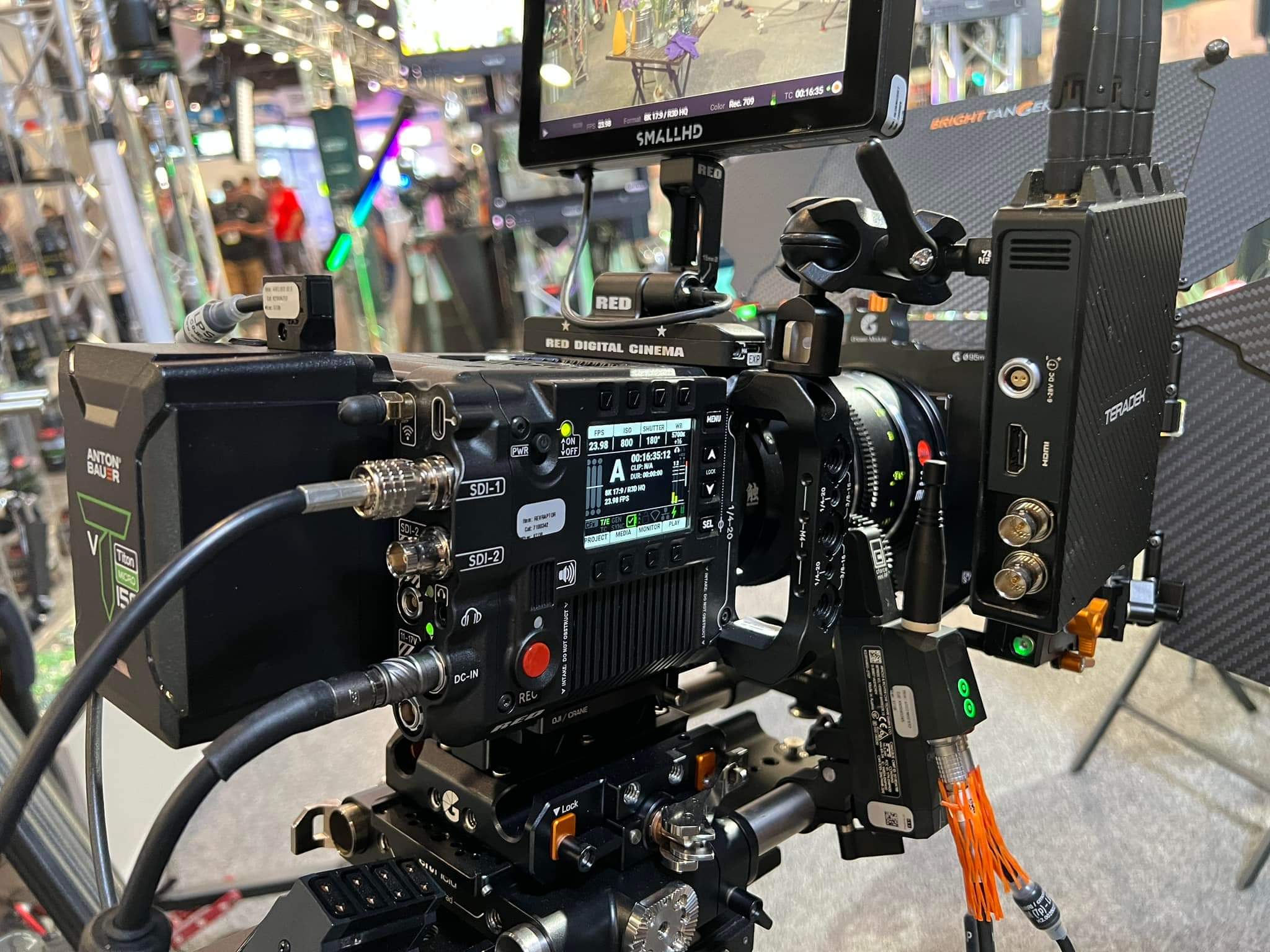 This image features a close-up view of a professional digital cinema camera, on a film or event set. The camera is equipped with various accessories and attachments. There are numerous cables connected to the camera, indicating it's set up for a high-quality live feed or recording. Attached to the top is a monitor. On the side, there's a battery pack providing a portable power source to the camera. To the right, there's a wireless network video streaming encoder with antennas for sending video to a remote monitor or director's station. The background is blurred, but we can discern a busy environment, likely a convention or trade show floor, suggesting this could be a demonstration or live event capture.
