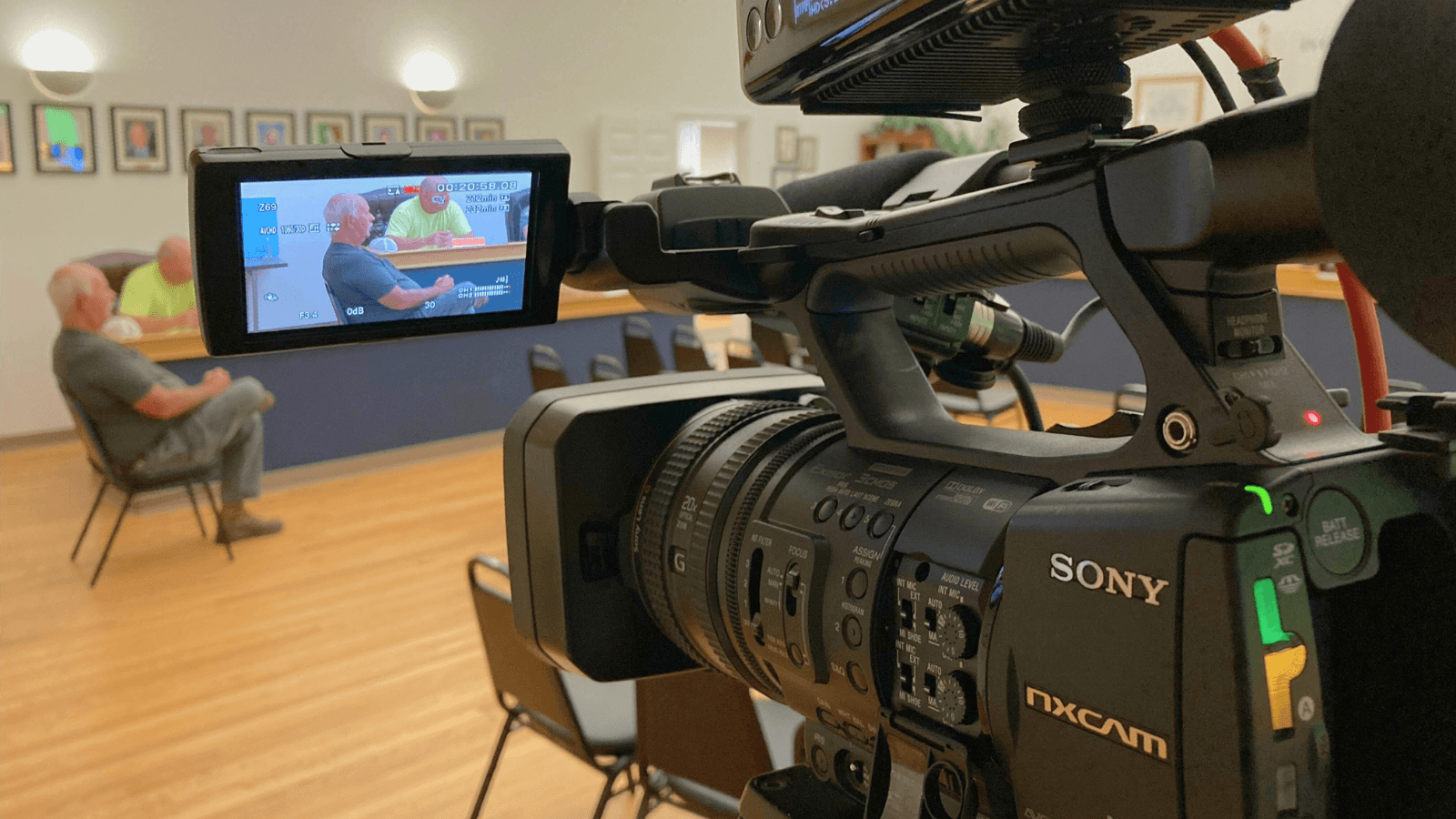 Close-up view of a Sony NXCAM video camera recording a meeting in a council chamber, with two men seated at a table visible on the camera's screen.