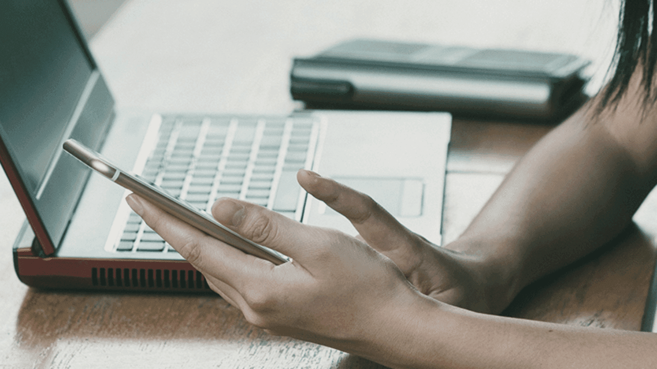 Hands hold a phone while laptop lays on desk demonstrating web usage