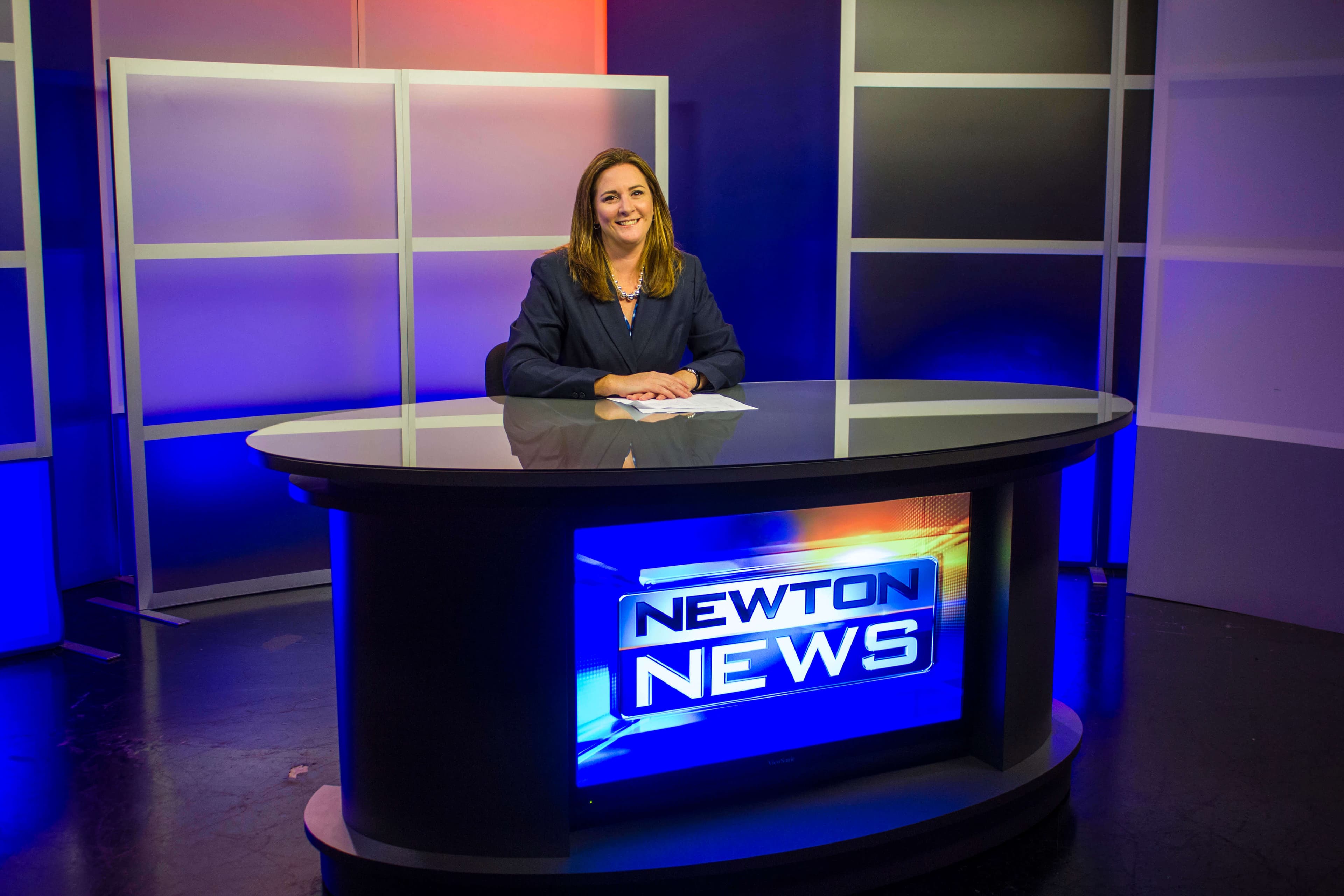 Community media station NewTV News Director Jenn Adams sits at news desk in a TV production studio for local news show Newton News
