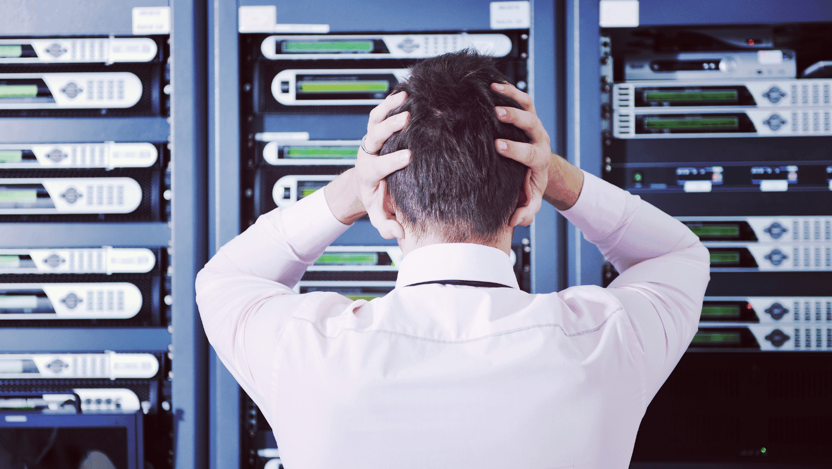 A man puts his hands on his head while stressfully staring at a wall with several servers