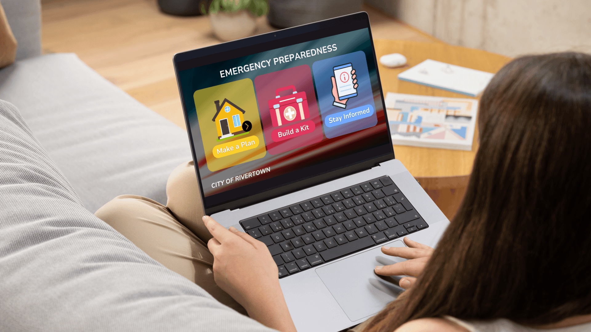 A person using a laptop displaying an emergency preparedness portal for the "City of Rivertown" with options to plan, build a kit, and stay informed.