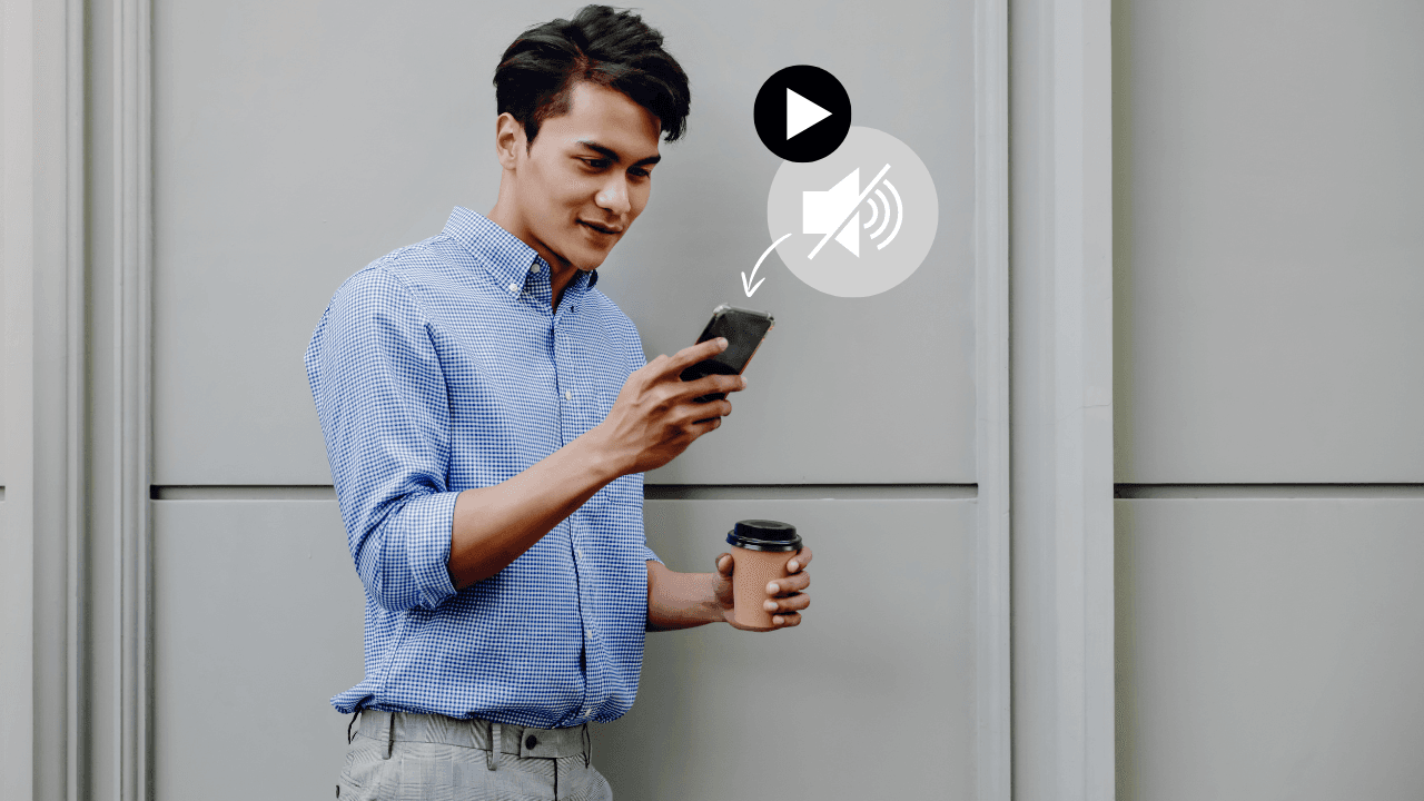 Man standing outdoors holding a smartphone and coffee cup, viewing content on his phone with a mute icon and play symbol indicating video playback without sound.
