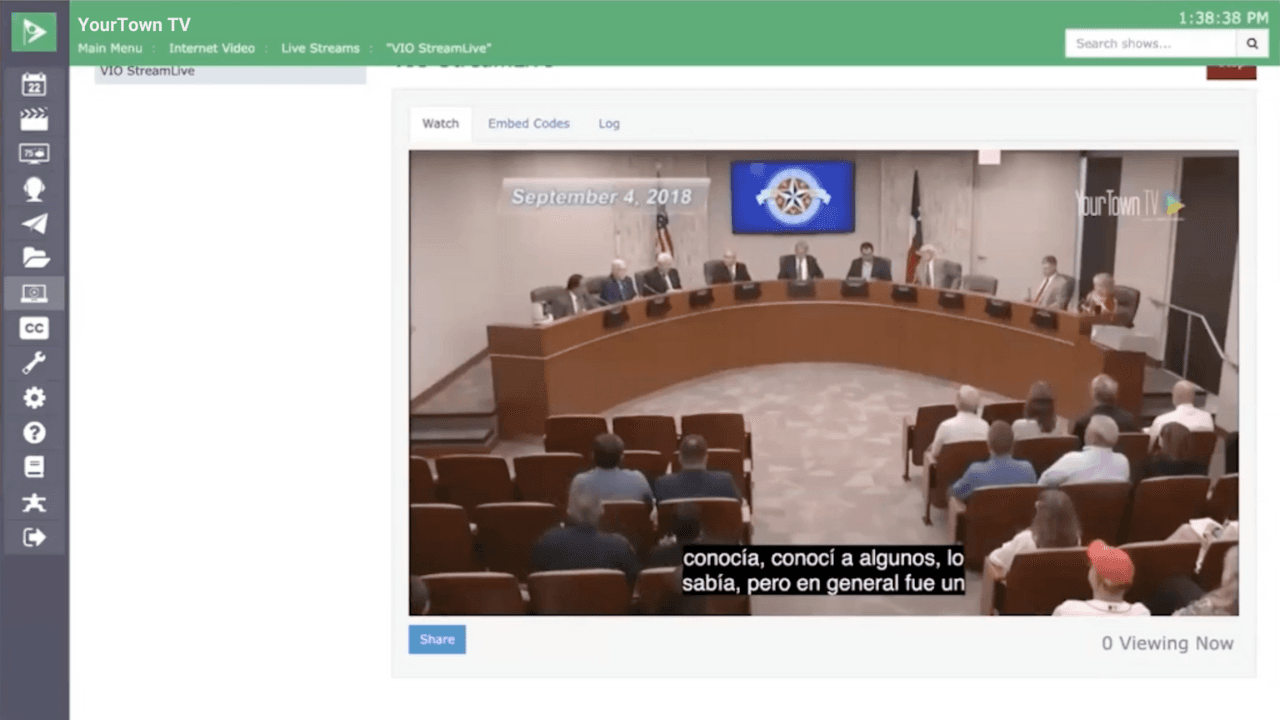 Alt text: Screenshot of a live stream of a government meeting being played in a Cablecast VIO interface. The stream shows a municipal council meeting with council members seated at a large semicircular table and an audience seated in rows. The video includes Spanish closed captions, demonstrating Cablecast Translate's real-time captioning capabilities. The interface displays navigation options, including menus for live streams, embedded codes, and logs, with a green header reading "YourTown TV" at the top.